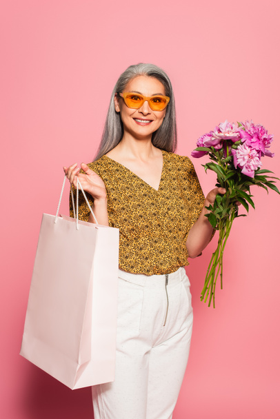 pleased asian woman in stylish sunglasses holding peonies and shopping bag on pink  - Fotoğraf, Görsel