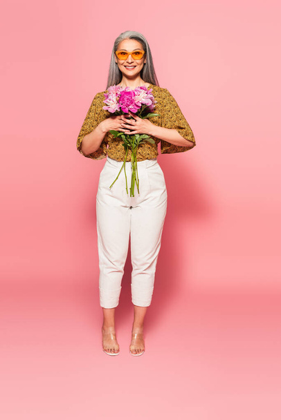 full length view of trendy asian woman with bouquet of peonies smiling at camera on pink  - Foto, Imagen