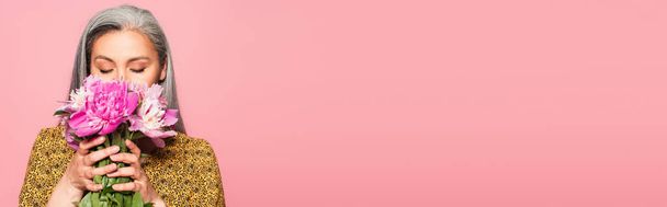 capelli grigi donna asiatica godendo sapore di peonie fresche con gli occhi chiusi isolati su rosa, striscione - Foto, immagini