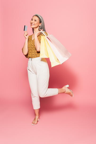 cheerful asian woman posing on one leg with shopping bags and credit card on pink background - Фото, изображение