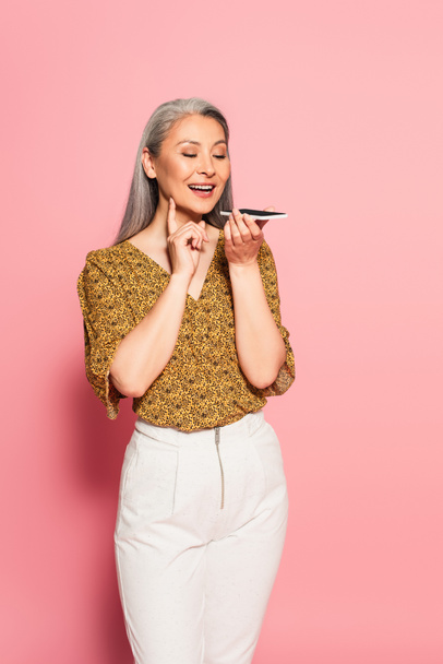 positive asian woman in trendy clothes sending voice message on pink background - Foto, imagen