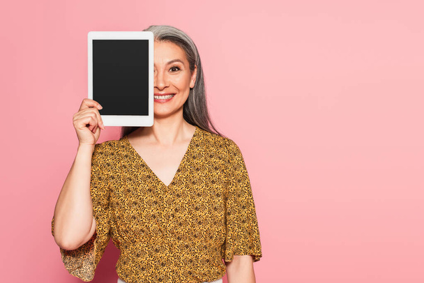 sonriente asiático mujer oscurecimiento cara con digital tablet aislado en rosa - Foto, imagen