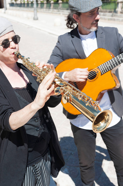 Μουσικοί τζαζ ντυμένοι με ρετρό στυλ παίζοντας στο δρόμο. Μια μελαχρινή γυναίκα με καπέλο και γυαλιά ηλίου που παίζει κλαρινέτο στο προσκήνιο και στο παρασκήνιο ένας άνδρας που παίζει κιθάρα. - Φωτογραφία, εικόνα