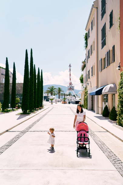 Annesi bebek arabasını şehir caddesinde yuvarlıyor ve küçük kız yakınlarda yürüyor. - Fotoğraf, Görsel
