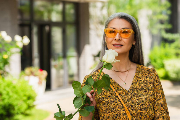 stylish asian woman in patterned blouse and sunglasses holding white rose outdoors - 写真・画像