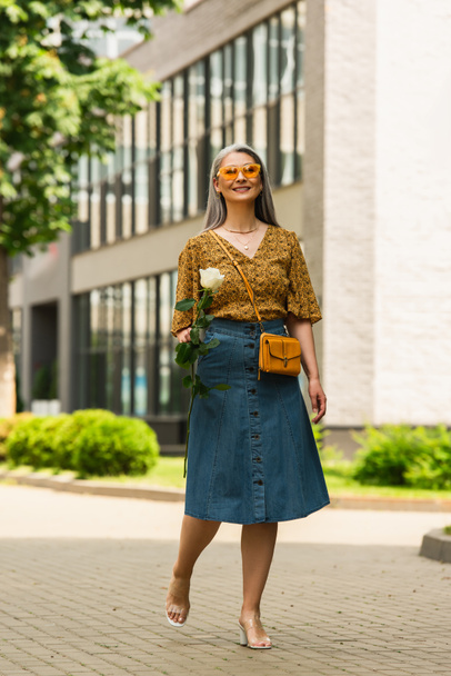 happy asian woman in denim skirt and patterned blouse walking with white rose outdoors - Фото, изображение