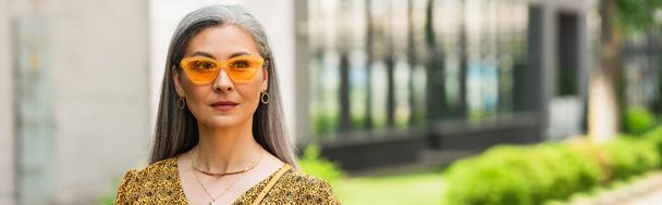 зрелая азиатская женщина в желтых солнцезащитных очках глядя в сторону на улице города, баннер - Фото, изображение