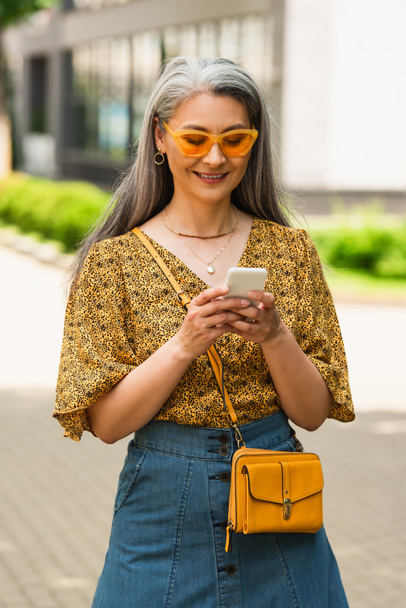 veselá asijské žena v slunečních brýlích a stylové oblečení psaní na mobilní telefon venku - Fotografie, Obrázek