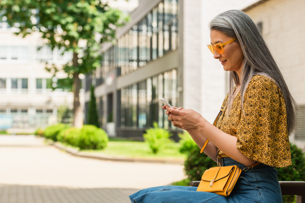 zralé asijské žena ve stylové sluneční brýle sedí na lavičce a zprávy na mobilním telefonu - Fotografie, Obrázek