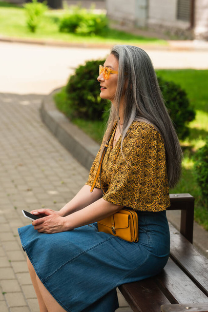 middle aged asian woman in trendy clothes sitting on city bench with smartphone - Foto, afbeelding