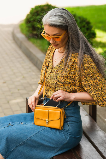 trendy asian woman with crossbody bag and smartphone sitting on bench in city - Fotografie, Obrázek