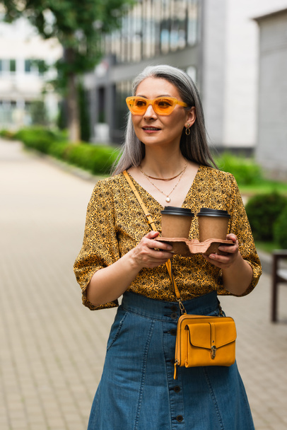 zralé asijské žena v vzorované halenky a žluté sluneční brýle stojící s kávou jít na ulici - Fotografie, Obrázek