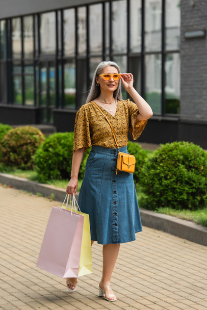 fashionable asian woman adjusting sunglasses while walking with shopping bags along city street - Photo, Image