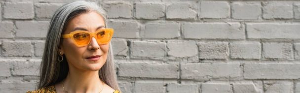 donna asiatica dai capelli grigi in occhiali da sole alla moda vicino al muro di mattoni all'aperto, banner - Foto, immagini