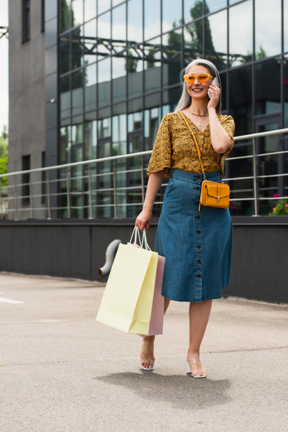 mature asian woman in trendy blouse and denim skirt talking on cellphone while holding shopping bags - Фото, зображення