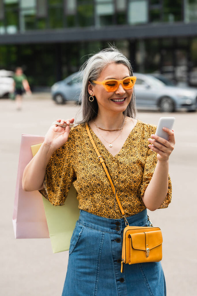 femme asiatique gaie en chemisier à motifs et lunettes de soleil en utilisant un téléphone portable tout en se tenant debout avec des sacs à provisions - Photo, image