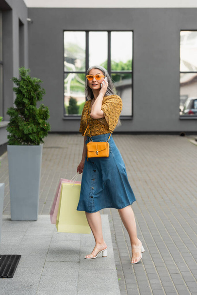 mujer asiática con estilo con bolsas de compras hablando en el teléfono móvil cerca del edificio - Foto, imagen