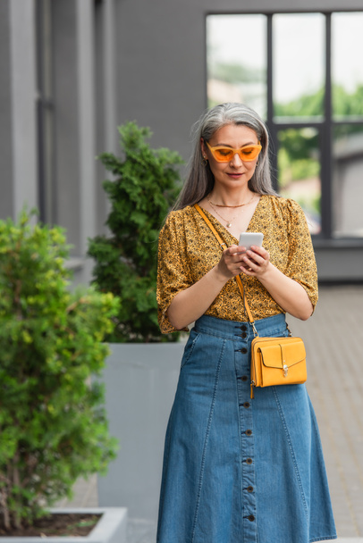 reife asiatische Frau in trendiger Kleidung und gelber Sonnenbrille auf dem Smartphone auf der Straße - Foto, Bild