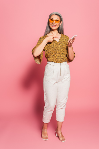 mature asian woman in stylish clothes and sunglasses pointing at smartphone on pink background - Valokuva, kuva