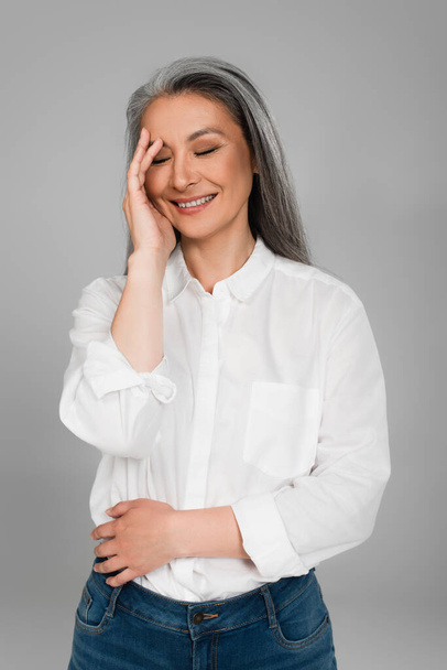 mature asian woman in white shirt touching face while laughing with closed eyes isolated on grey - Foto, Bild