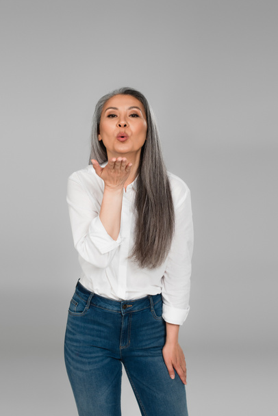 middle aged asian woman in white shirt and blue jeans blowing kiss at camera isolated on grey - 写真・画像