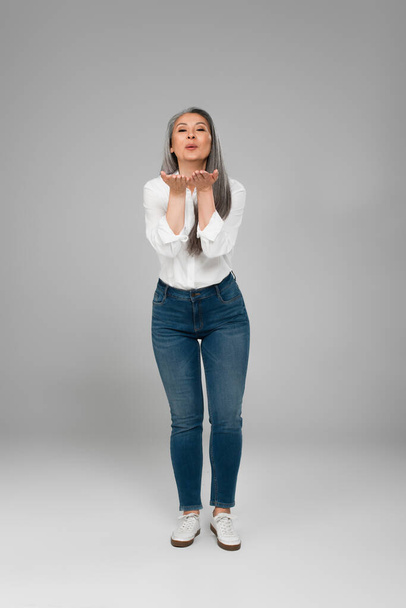full length view of asian woman in jeans and white shirt blowing air kiss on grey  - Foto, Imagem