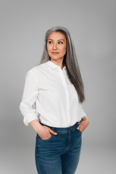 middle aged asian woman standing with hands in pockets of jeans isolated on grey - Фото, изображение