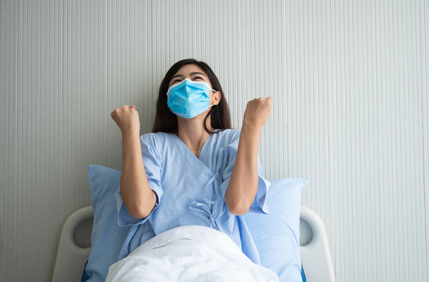 Ευτυχισμένη Ασιάτισσα ασθενής με μάσκα, ξαπλωμένη στο κρεβάτι, σήκωσα τα χέρια μου και χάρηκα που έδειξα εμπιστοσύνη στη θεραπεία. Έννοια της πίστης στη θεραπεία και ασφαλιστική κάλυψη - Φωτογραφία, εικόνα