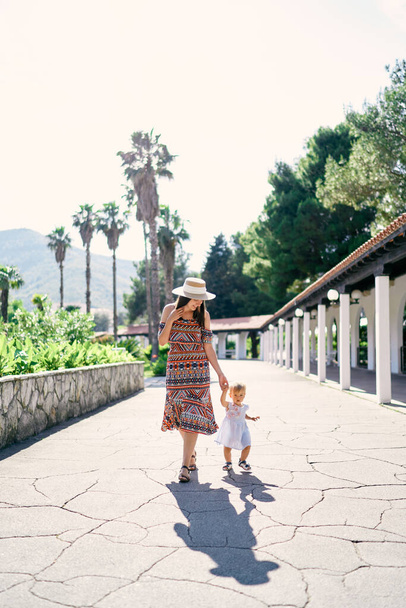 Η μαμά περπατά με ένα κοριτσάκι σε ένα πλακόστρωτο μονοπάτι στο πάρκο με φόντο φοίνικες και πράσινο - Φωτογραφία, εικόνα