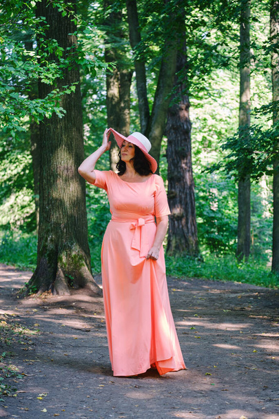 beautiful adult woman in a long coral dress and a wide hat walks in a summer par - Fotoğraf, Görsel