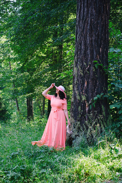 beautiful adult woman in a long coral dress and a wide hat walks in a summer par - Photo, image