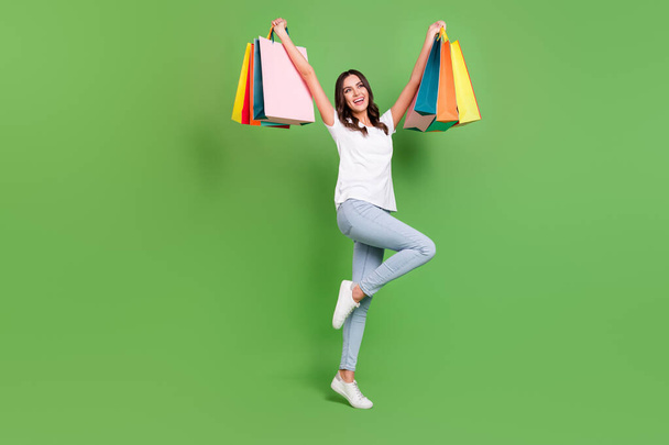 Full body photo of cool brunette millennial lady hold bags wear t-shirt jeans isolated on green color background - Фото, зображення