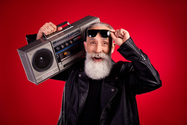 Photo of elderly man happy positive smile hold retro boombox sound rocker hand touch glasses isolated over red color background - Fotografie, Obrázek