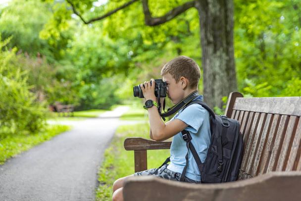 Турист с фотоаппаратом. Мальчик с фотоаппаратом фотографирует природу в парке. Молодой фотограф сидит на скамейке в парке и фотографирует природу. Путешествие с камерой - Фото, изображение