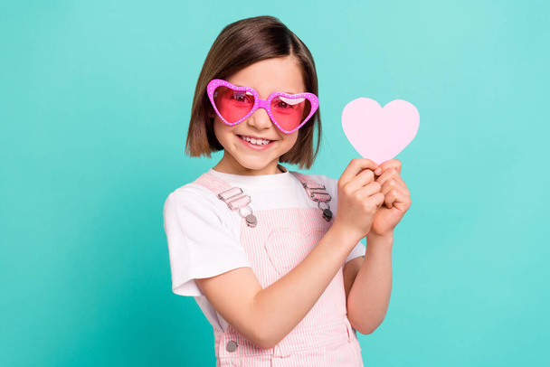 Photo of funny adorable school girl wear pink overall glasses smiling holding little heart isolated teal color background - Фото, изображение