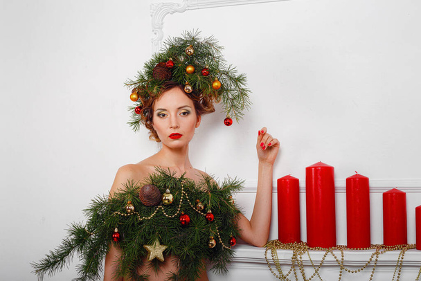 Красивая счастливая молодая леди с рождественским сосновым венком на голове и модным платьем из елки, украшенной рождественскими шарами, ягодами и снежинками. Новогодняя концепция - Фото, изображение