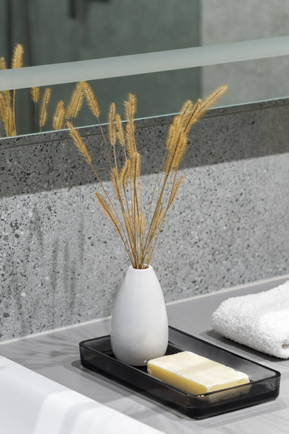 Close-up on decorative white vase with dried grass and natural soap bar on tray next to white towel in bathroom with granite tiles - Foto, imagen