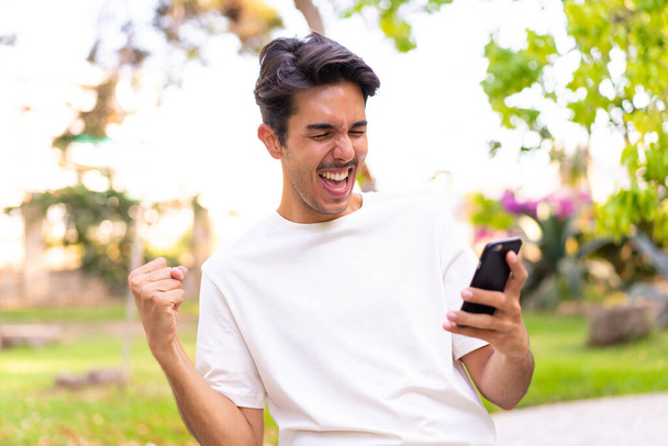 Young caucasian man in a park using mobile phone and doing victory gesture - Foto, Imagen