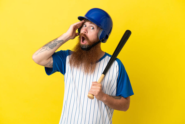 Redhead baseball player man with helmet and bat isolated on yellow background doing surprise gesture while looking to the side - Foto, Imagem
