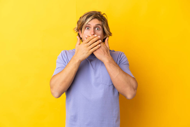Knappe blonde man geïsoleerd op gele achtergrond bedekken mond met handen - Foto, afbeelding
