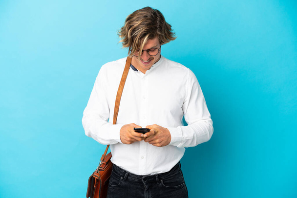 Молодий блондинка бізнесмен ізольований на синьому фоні, відправляючи повідомлення з мобільним
 - Фото, зображення