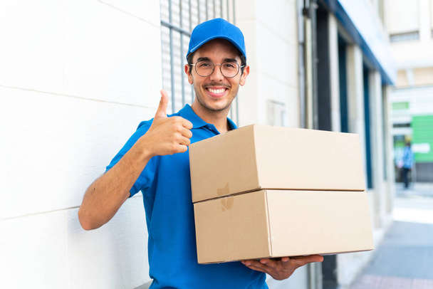 Молодой доставщик на улице держит коробки с большим пальцем вверх - Фото, изображение