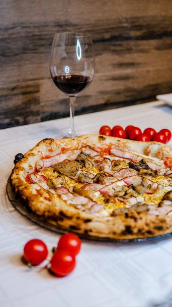 A delicious pizza with ham and cherry tomatoes and a glass of red wine at a restaurant - Foto, afbeelding
