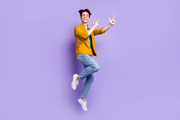 Full size photo of cool brunet millennial guy jump point empty space wear shirt jeans isolated on violet color background - Photo, Image