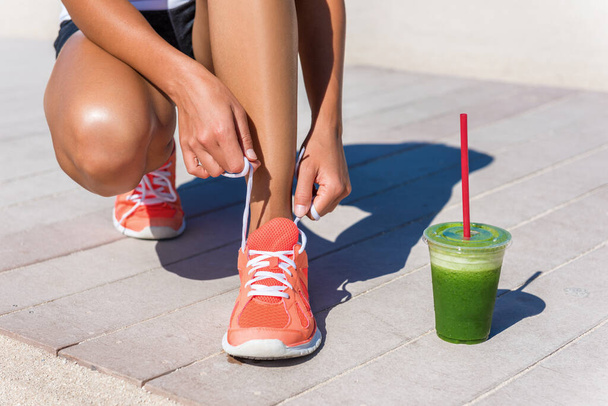 Running woman athlete runner getting ready for beach morning run by tying shoe laces of running shoes with green vegetable smoothie breakfast. Closeup on feet. Fitness and healthy lifestyle concept - Foto, Imagem