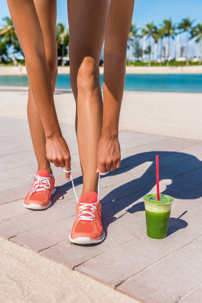 Running woman runner getting ready for workout by tying shoe laces of running shoes with green vegetable smoothie on beach boardwalk. Closeup of legs. Fitness and healthy lifestyle concept. - 写真・画像