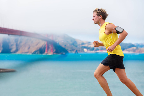 Runner man listening to music during run cardio workout. Running athlete using app on smartphone armband and earphones. Sporty fit young jogger jogging by San Francisco Bay and Golden Gate Bridge. - Foto, imagen