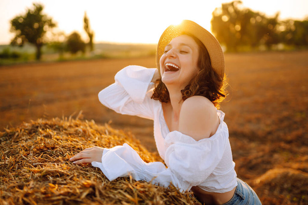  Beautiful curly woman in hat and clothes posing near hay bales in the countryside at sunset. Fashion concept. Nature, vacation, relax and lifestyle. - Φωτογραφία, εικόνα