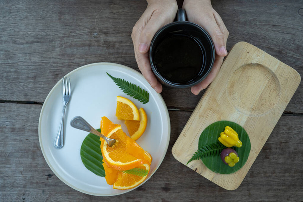 A hand holding a mug of black coffee with plates of fresh fruits next to it on a wooden table - Foto, imagen