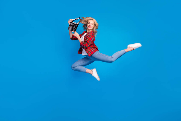 Photo of adorable funky young lady wear plaid shirt smiling humping high holding clapperboard isolated blue color background - Foto, immagini
